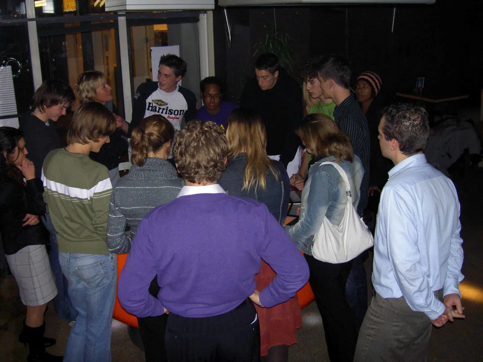 A crowd of a dozen people standing shoulder to shoulder around the orange prototype, playing.