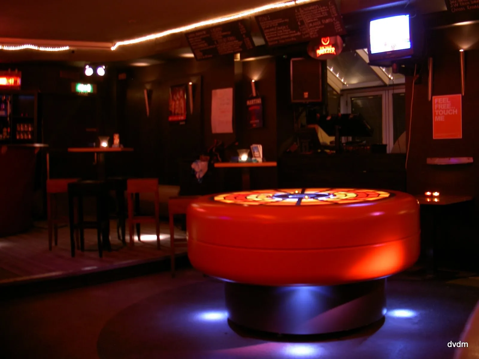 The table alone in a bar before anyone has arrived, glowing on a dark floor among stools and high tables, a poster on the wall behind it reading "Feel free... touch me".