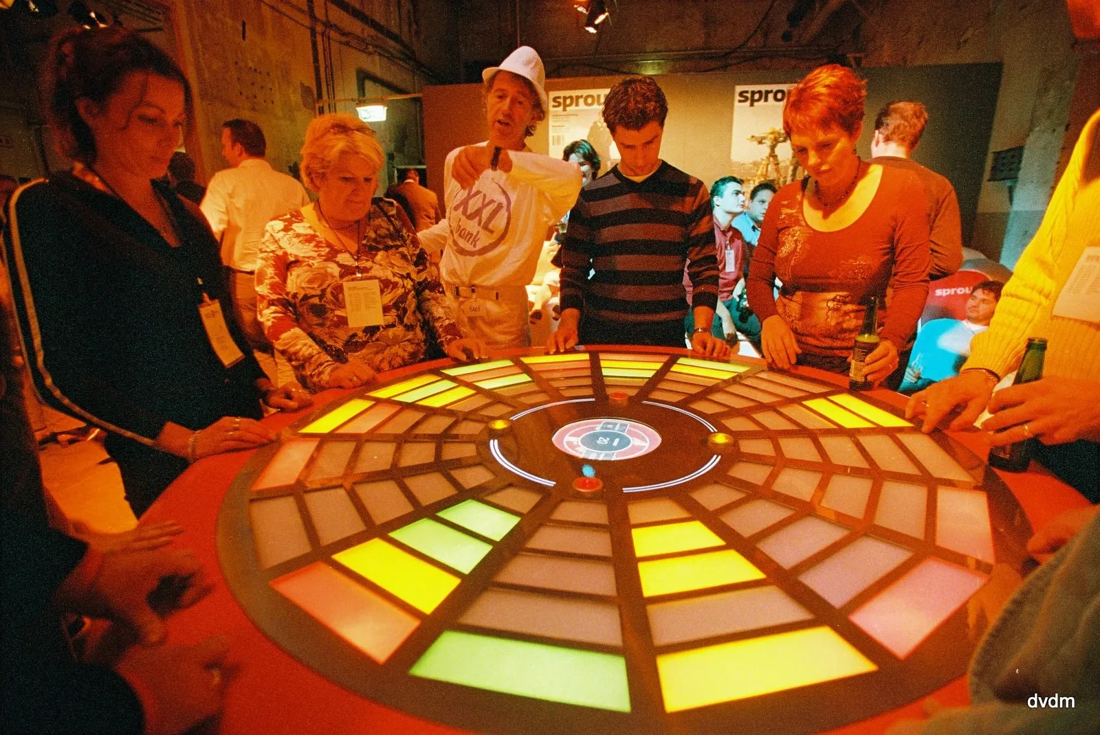 Half a dozen people crowded round the lit table at an event, hands flat on the field and beer bottles in hand, with banners and a bare brick hall behind them.