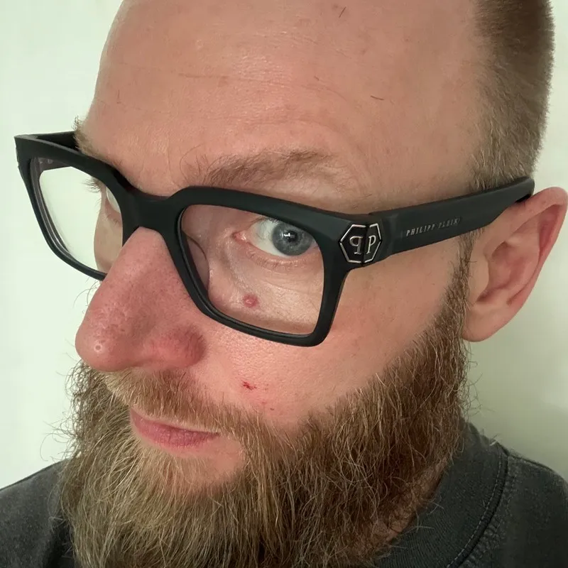 Dirk van de Mortel, close up in dark-framed glasses against a plain wall.