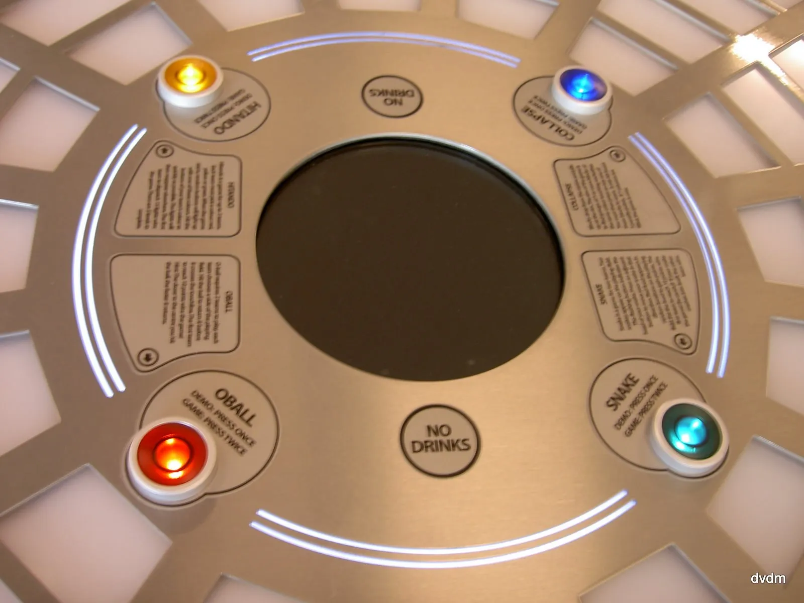 The centre of the table: four lit start buttons in amber, blue, red and green, each naming its game (Hitando, Collapse, Oball, Snake) and reading "demo: press once, game: press twice", with the rules of all four engraved into the metal ring around the screen.
