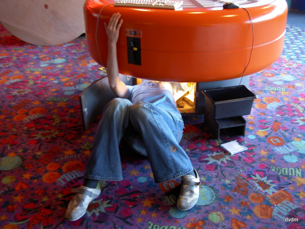 A man lying on his back on a brightly patterned arcade carpet, legs bent and trainers
    on the floor, with his head and shoulders inside the open base of a round orange gaming
    table and one hand braced against its side.
