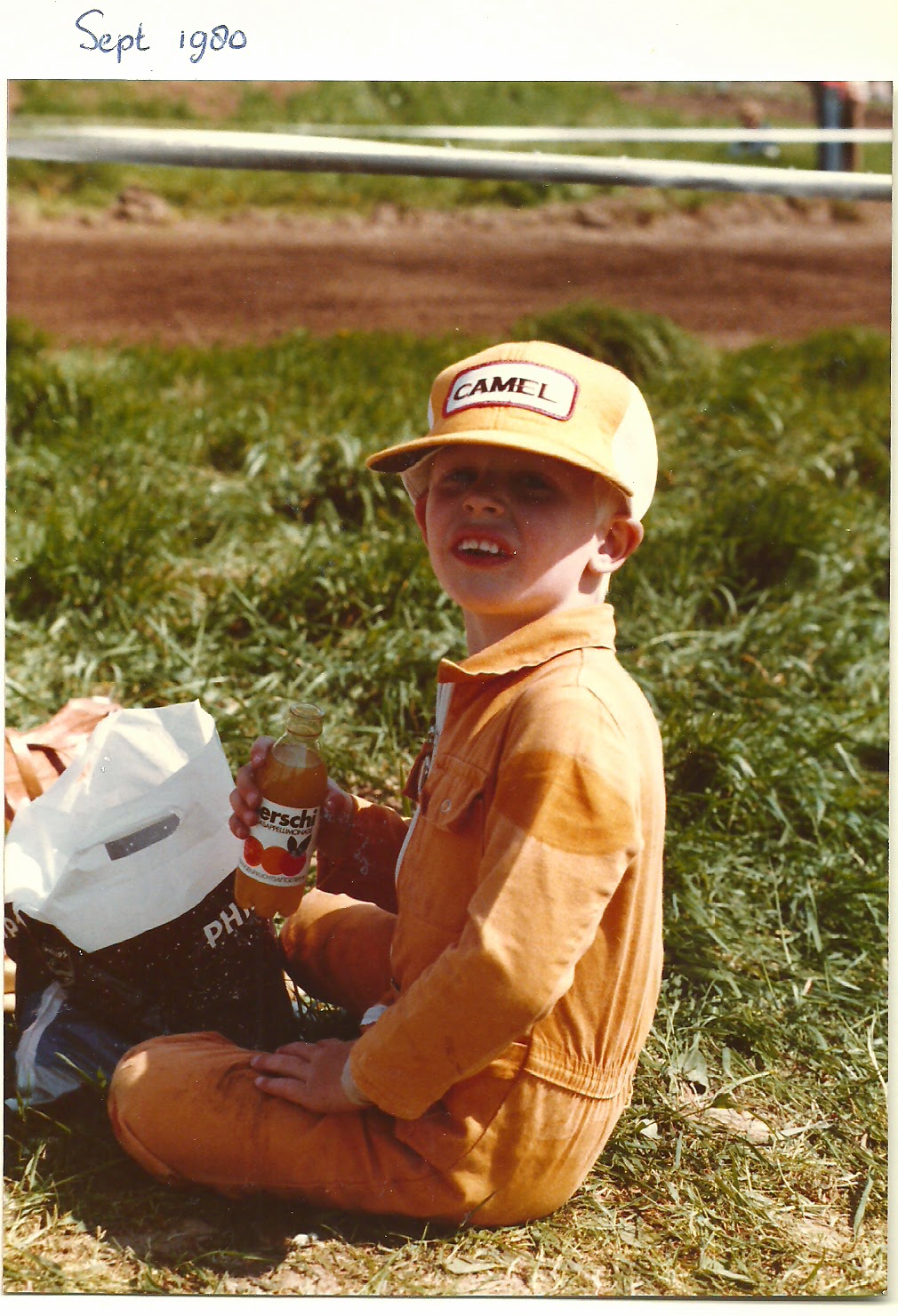 A small boy in orange overalls and a Camel cap sits cross-legged on the grass beside a
      motocross track, holding a bottle of orangeade, with the rutted dirt course behind him.